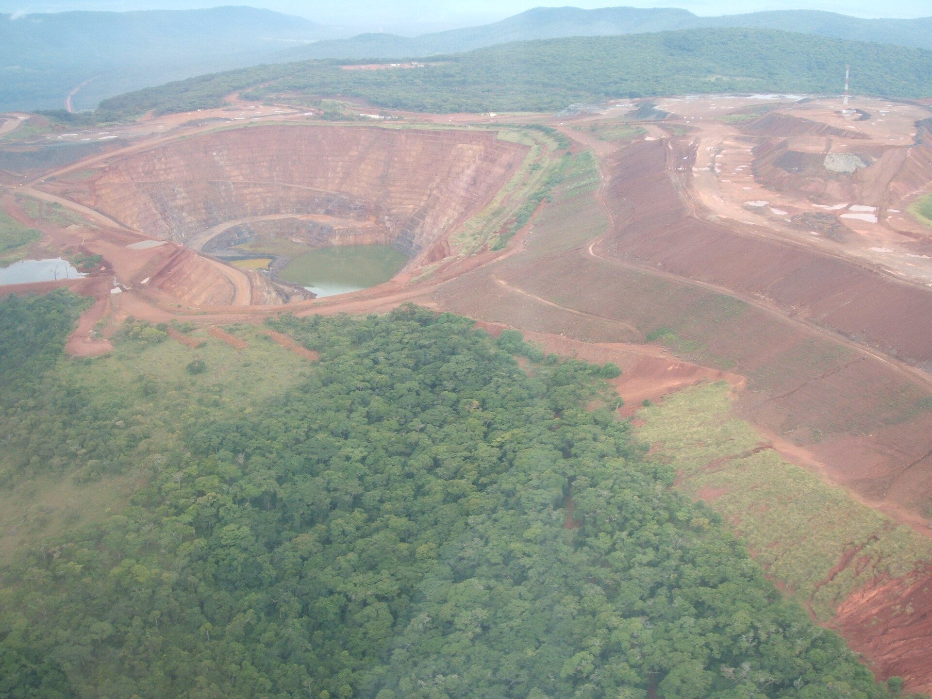 An aerial view of a large open-pit gold mine in Tanzania, its terraced red walls curving down to a small green pool, with forest and hills around it.