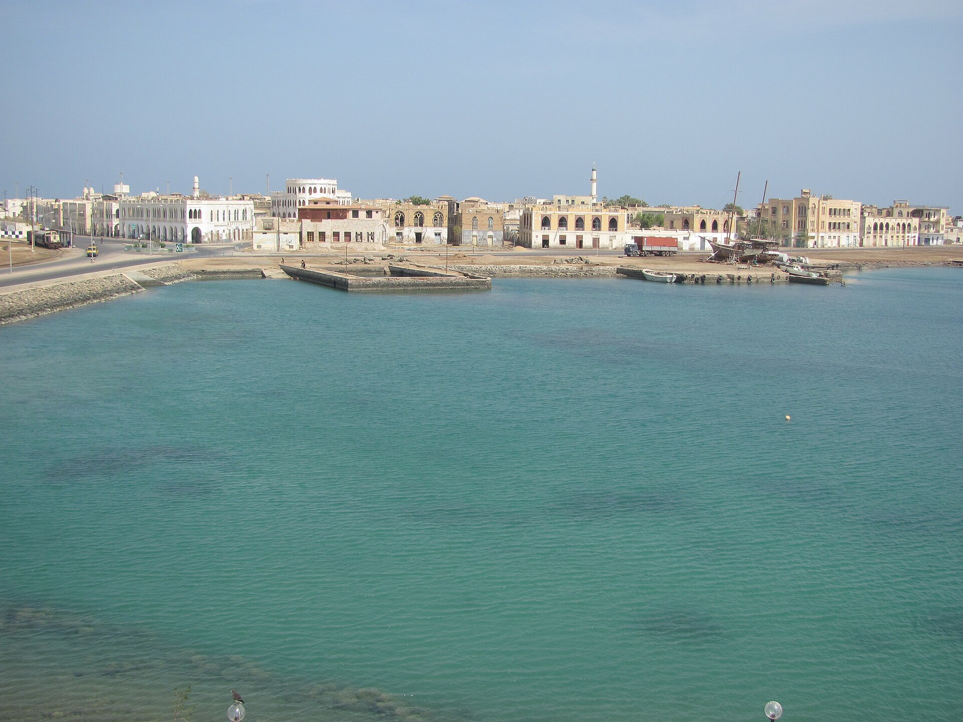 Low stone buildings along the waterfront of an old port town, with a stone quay, a few wooden boats and shallow turquoise water in the foreground.