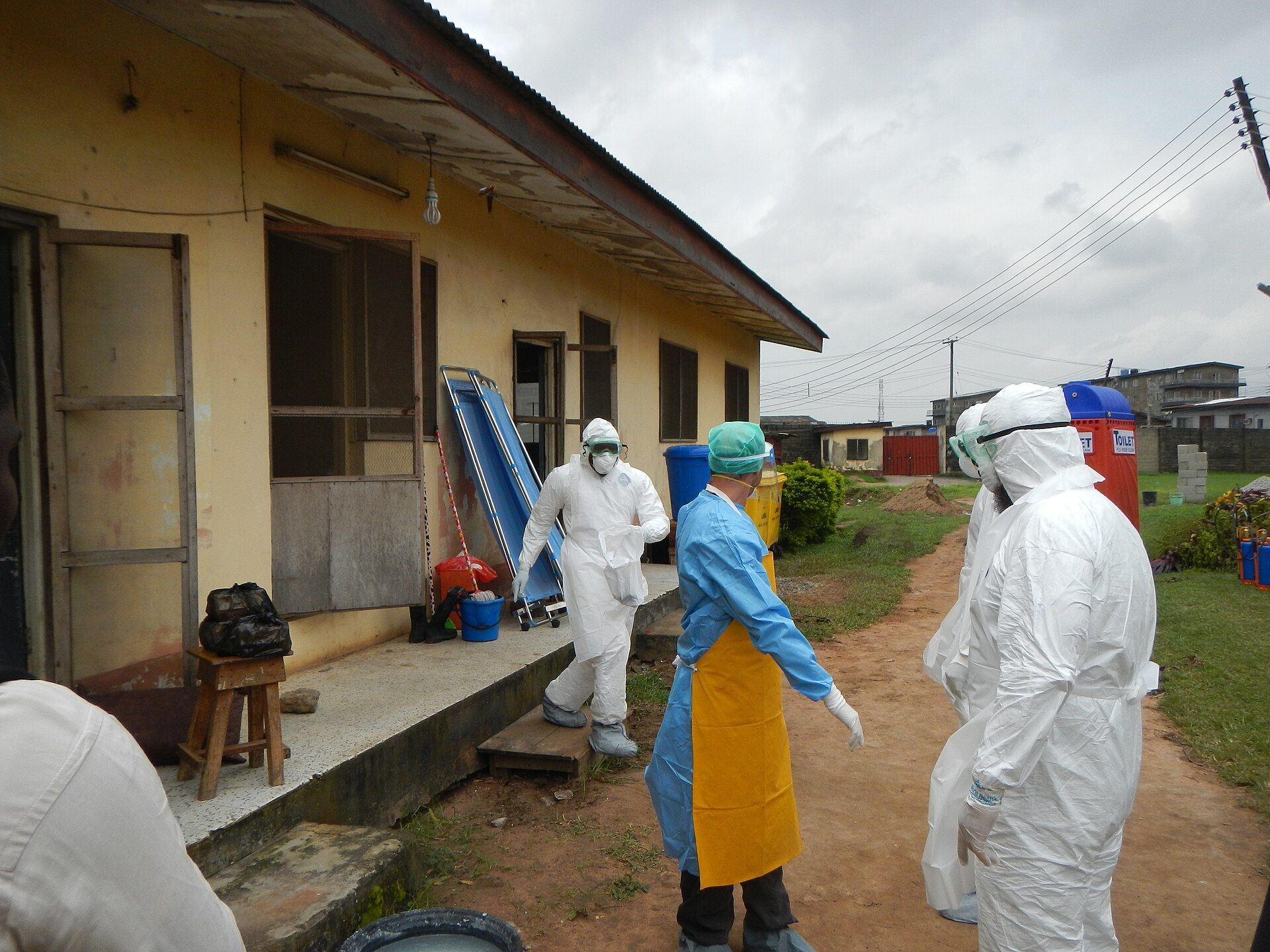 Three health workers in white protective suits, masks and goggles, and one in a blue gown and yellow apron, stand outside a low yellow building in Lagos as they get ready to enter an Ebola isolation ward.