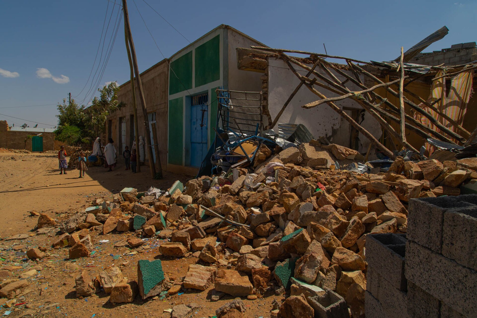 A collapsed house in a street in Hawzen, Tigray, with a pile of rubble and broken roof beams. A few people stand far down the street.