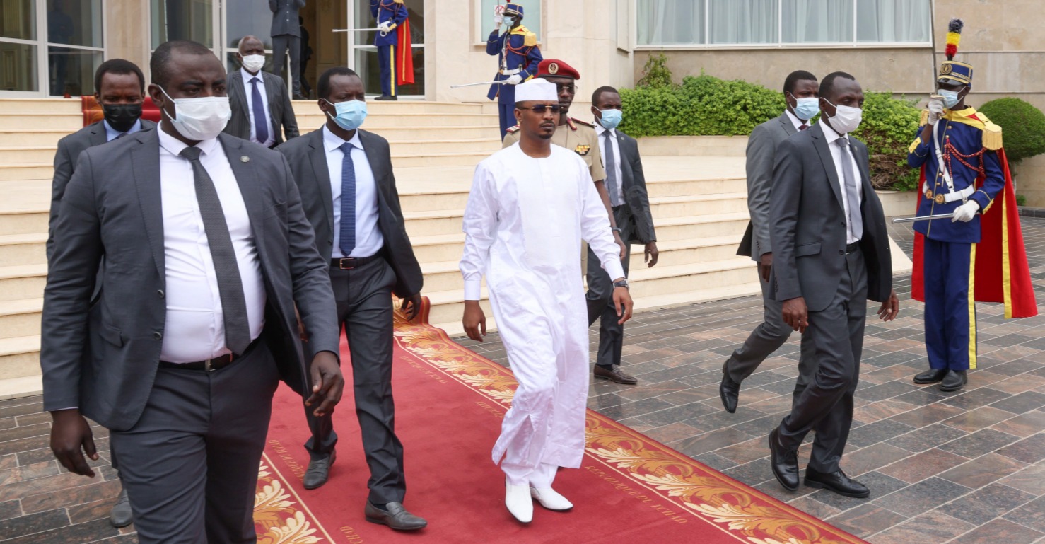 Chad's president, in a white robe and cap and dark glasses, walks on a red carpet outside a palace, followed by men in grey suits and face masks, with ceremonial guards in blue uniforms and red capes standing at the edges.