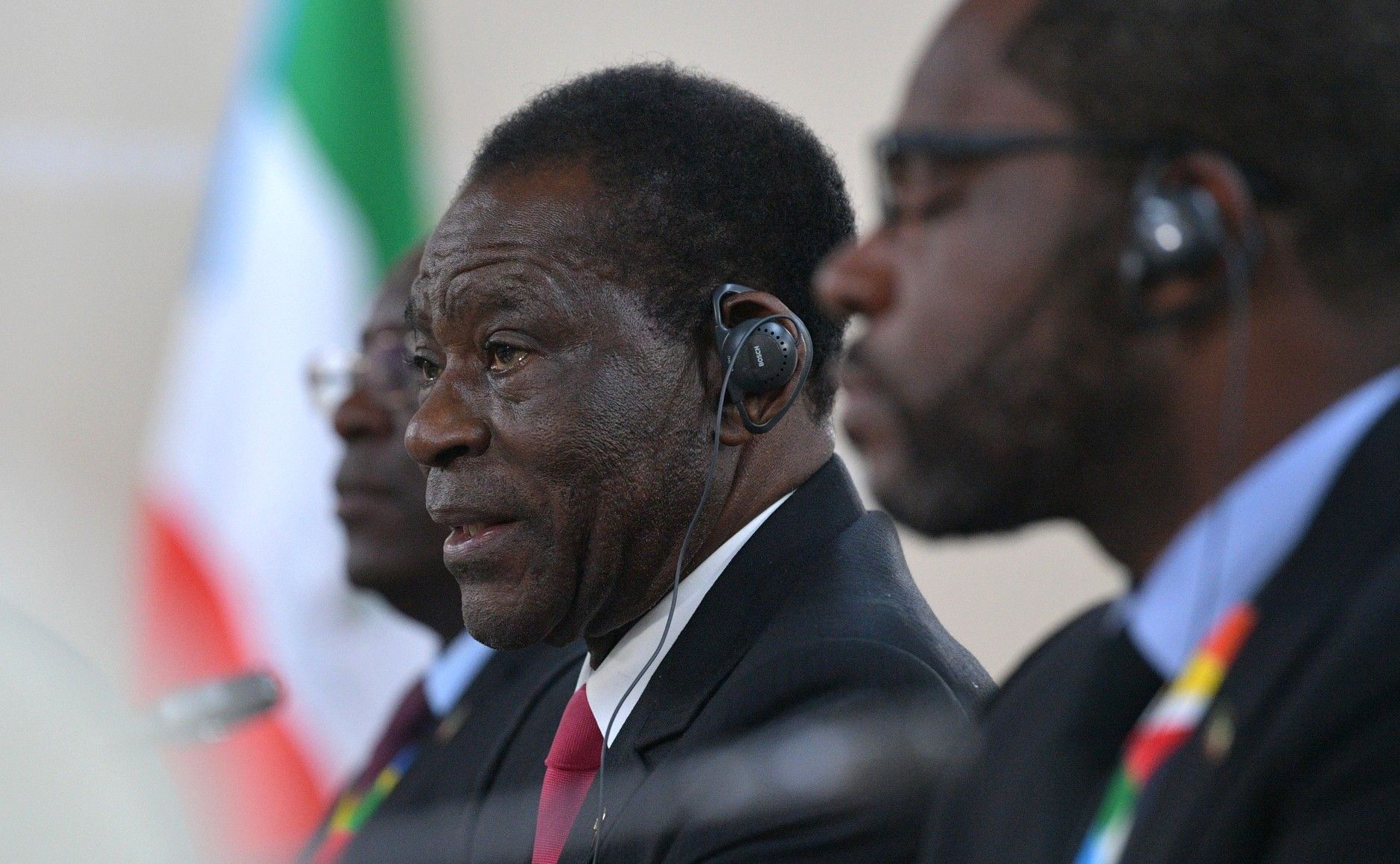 Equatorial Guinea's president in profile in a dark suit and red tie, wearing a translator's earpiece, with the country's green, white and red flag blurred behind him and an out-of-focus man in glasses on each side.