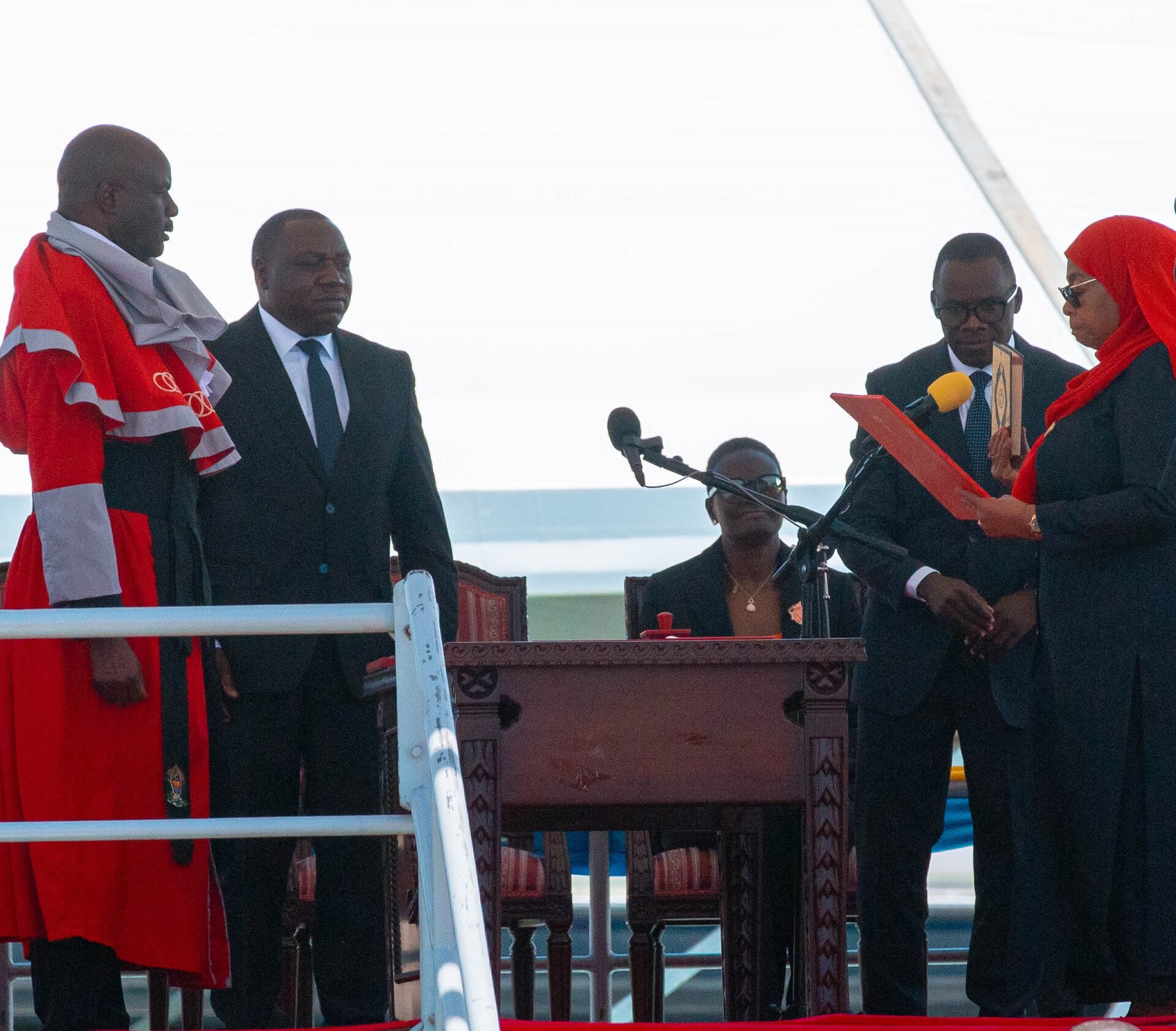 A woman in a red headscarf and dark clothes, at the right, holds a document and a book while taking the oath of office, facing a judge in a red robe at the left, with a microphone and a wooden table between them and other officials standing nearby.