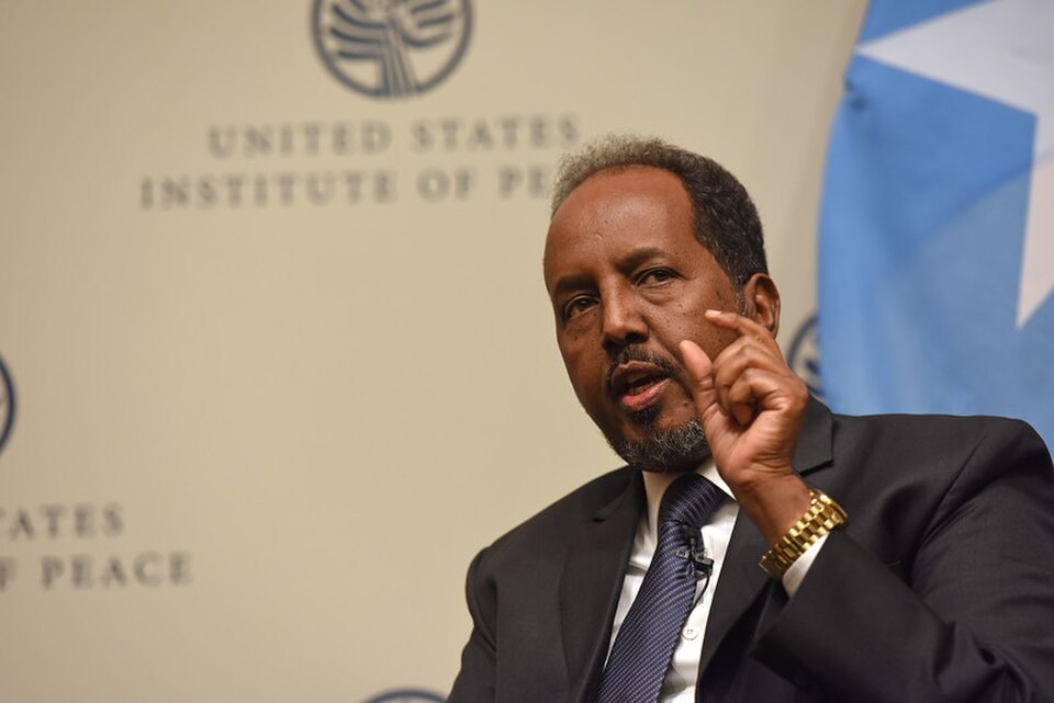 Hassan Sheikh Mohamud, in a dark suit and blue tie, raises one finger as he speaks, with a Somali flag behind him and a United States Institute of Peace backdrop.