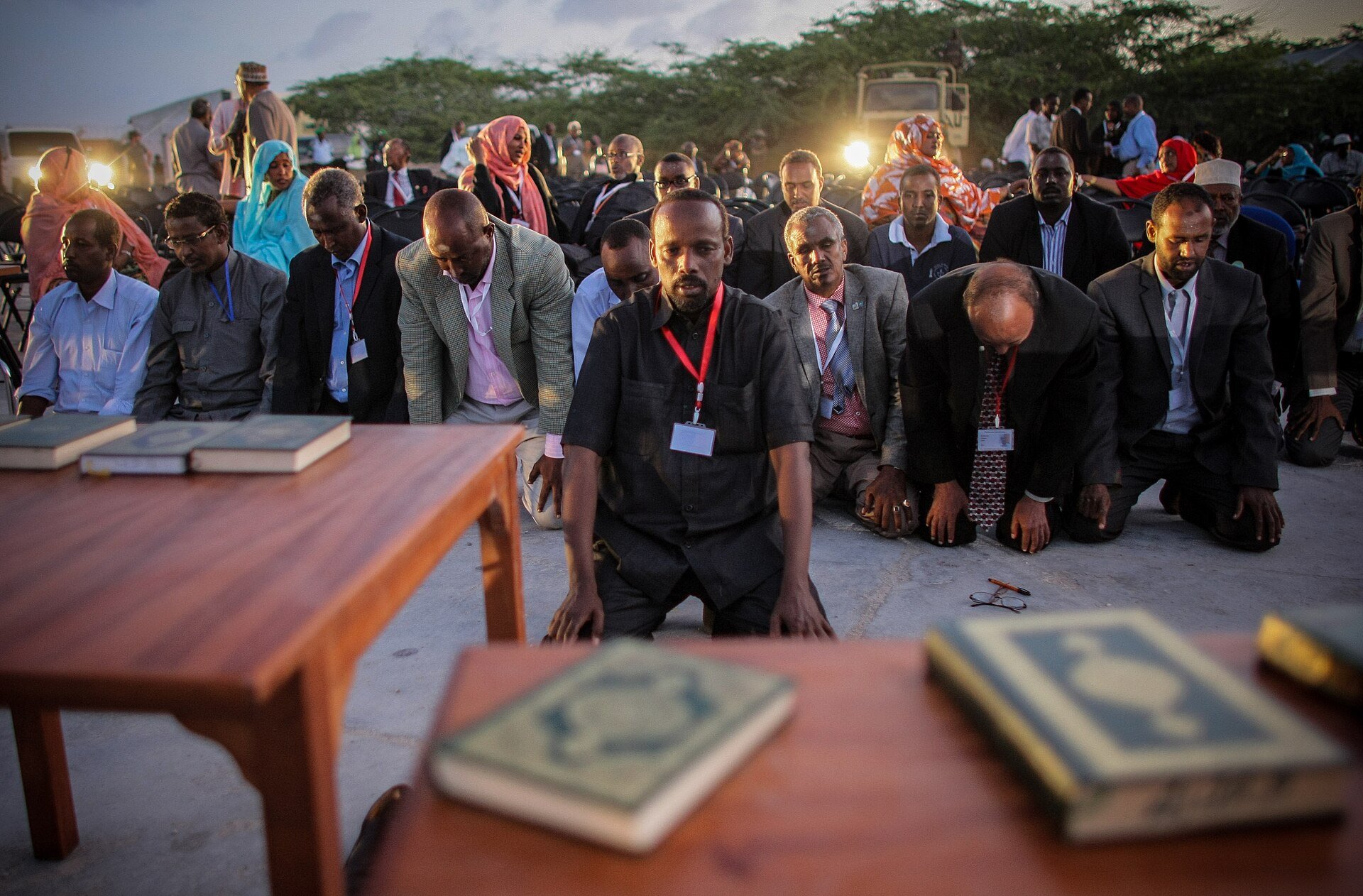 Newly elected Somali members of parliament kneel in rows in prayer at dusk, some wearing red lanyards, behind tables holding copies of the Quran.