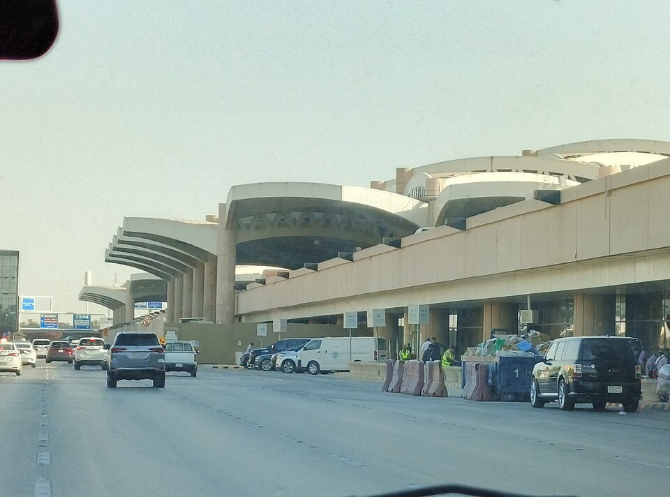 A busy road beside the long, curved concrete terminal building at King Khalid International Airport in Riyadh, with cars on the road, workers beside a dumpster and a hazy sky above.