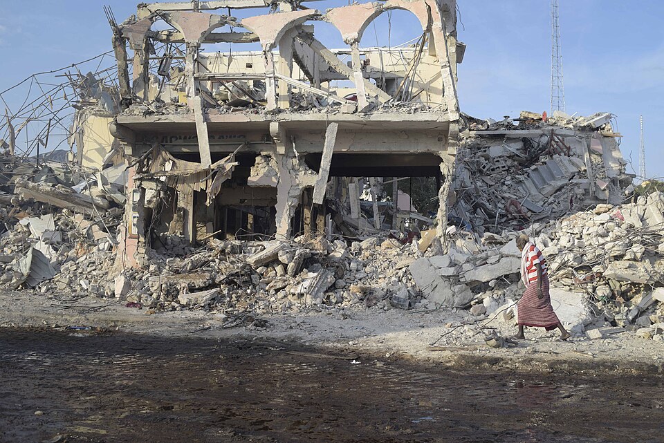 An elderly man in a striped top and a long wrap skirt walks past the collapsed, gutted front of a multi-storey building and piles of rubble under a blue sky.
