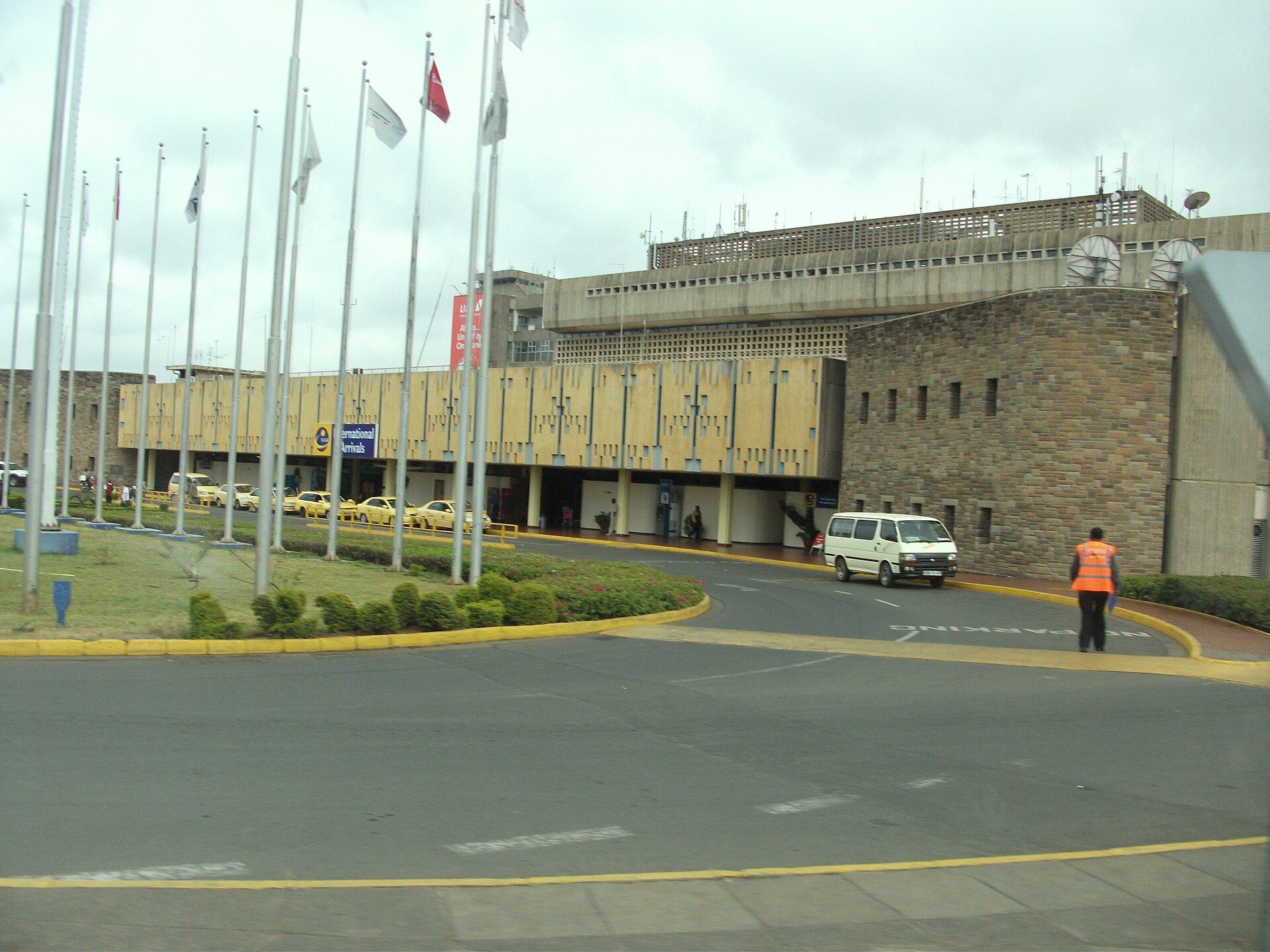 A low brown stone terminal building with a long canopy, flagpoles in front, a white minibus and taxis on the forecourt, and a man in an orange vest walking across the road.