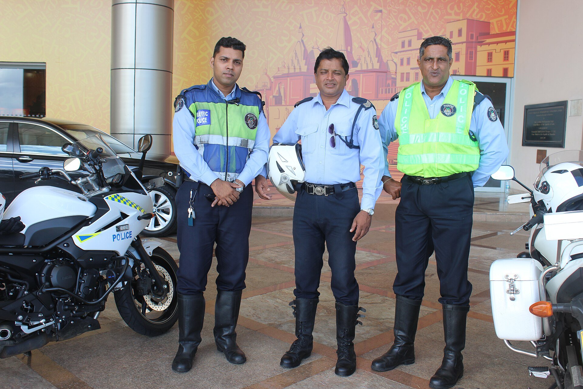 Three Mauritian police officers in light-blue shirts and dark trousers stand side by side outside a building with an orange mural, two of them in high-visibility vests, one marked Traffic Police, with white police motorbikes at each side.