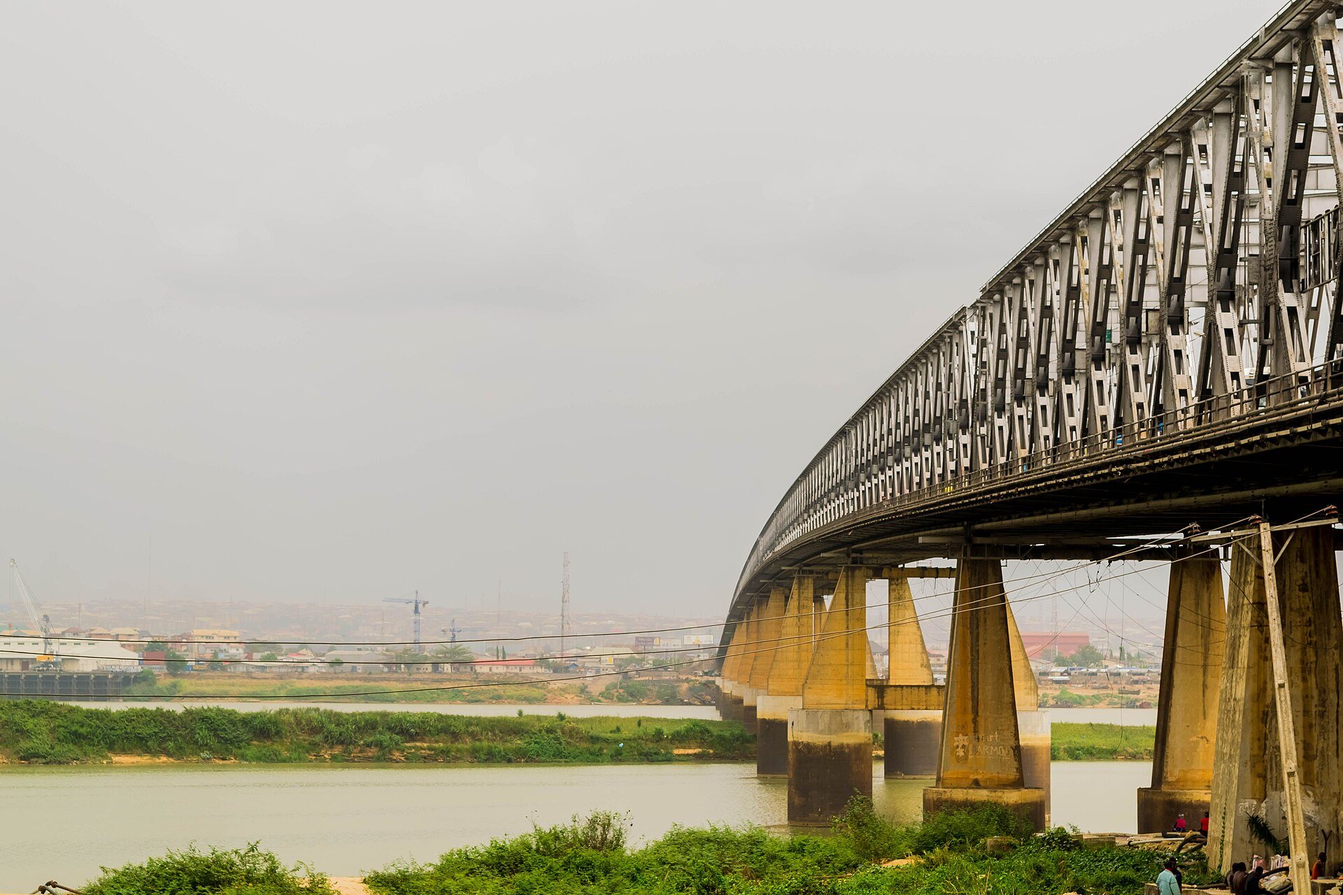 The Niger Bridge at Onitsha curving along the river on tall concrete pillars, with the town on the far bank under a grey sky.