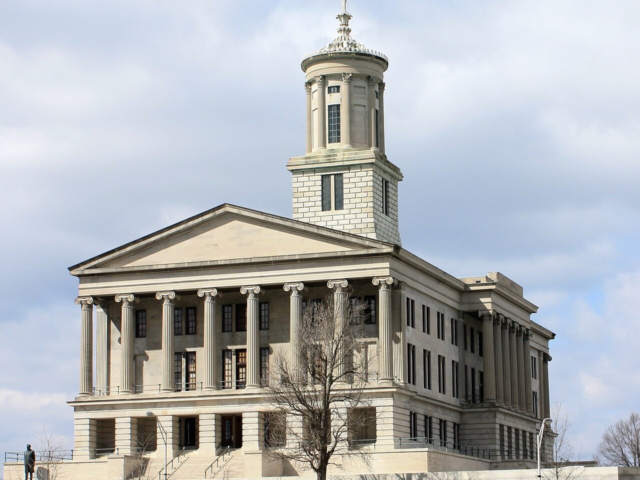 The Tennessee State Capitol in Nashville: a pale stone building with a columned front and a square tower topped by a small round cupola, seen from below under a cloudy sky.