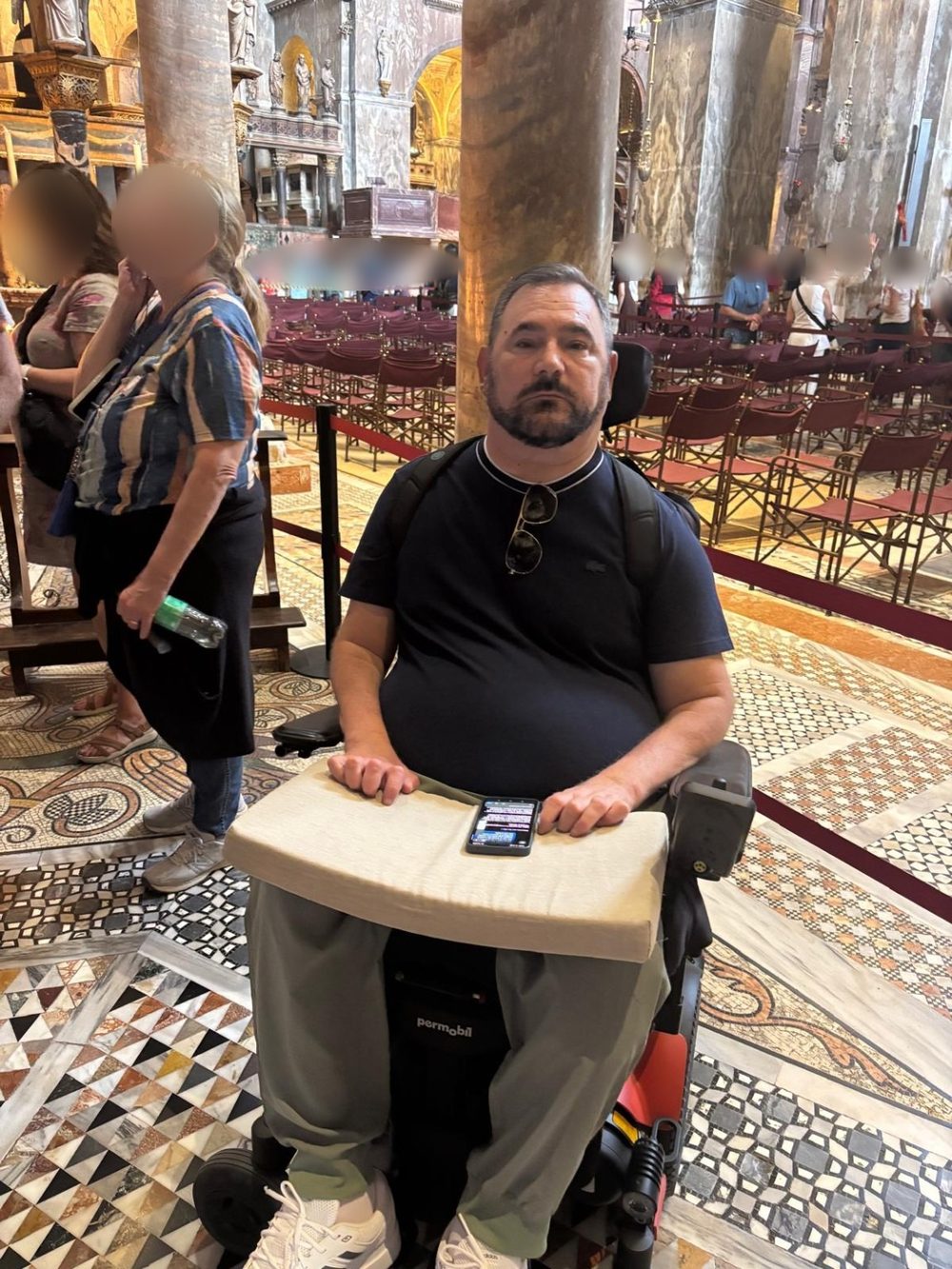 A man with a short dark beard sits in a powered wheelchair with a cushioned tray across his lap, looking at the camera, inside a large Basilica with marble columns, a patterned mosaic floor and rows of red chairs behind him.